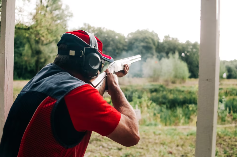 man in red and black clothing and hat holding a gun and shooting,the field with grass and trees